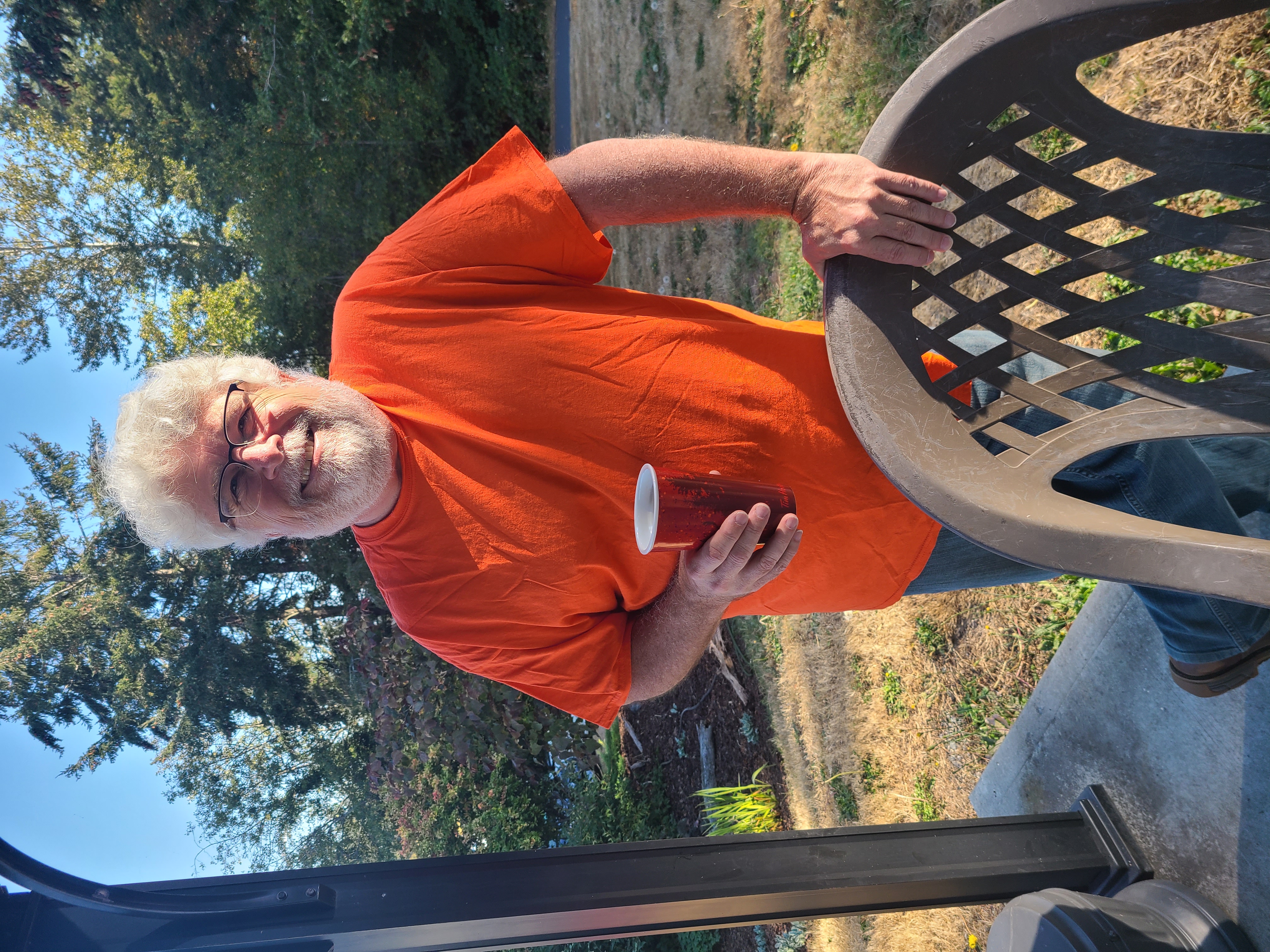 A Photo of Richard Gale in an orange shirt, holding a red cup in one hand while resting the other against a chair.