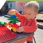 A toddler builds a tower with block at an activity table outdoors in the sunshine. 