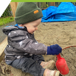 A baby dressed in Muddy Buddies, mitts and a hat is happily digging in a sandbox.  She is covered in sand and laughing.