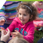 A litttle girl around three years old is sitting in a circle with her friends playing a game. 