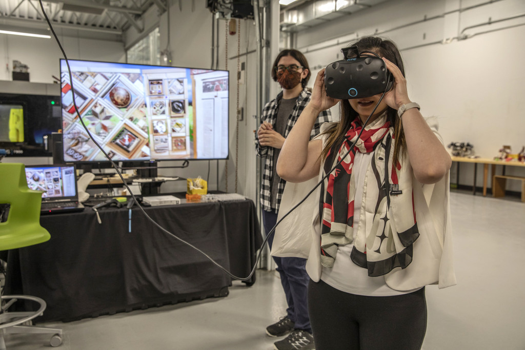 A woman in the foreground wears a VR head set. A man stands next to a large monitor with images on it in the background.