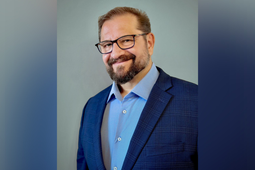 Portrait style photograph of a man with a goatee and glasses smiling at the camera. He is a wearing a bright blue sports coat and open collar, light blue dress short.