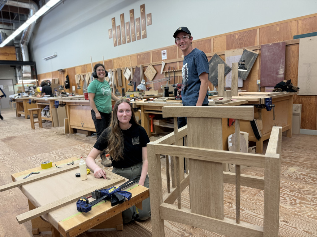 Three students - two female and one male - are in a fine furniture classroom. One student is kneeling and the other two are standing. It looks like they have stopped work on projects to smile and pose for a photograph.