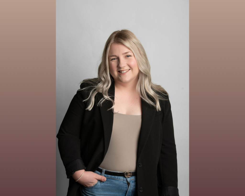 A woman with long blonde hair, a black jacket and brown shirt stands in front of a grey background.