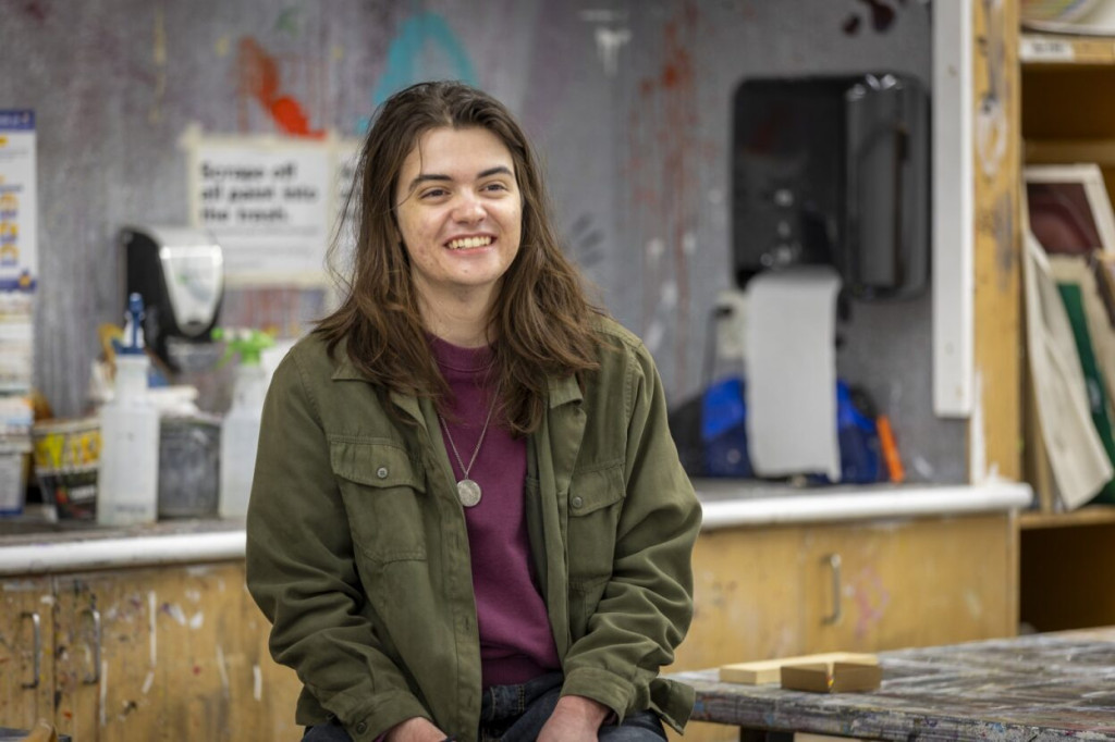 A guy with long hair smiling, while sitting in a visual arts classroom.