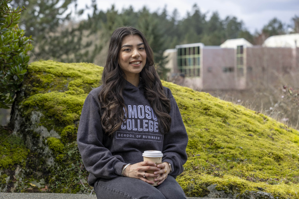 Sabrina sitting outdoors, wearing a Camosun hoodie, holding a cup of coffee, and smiling.