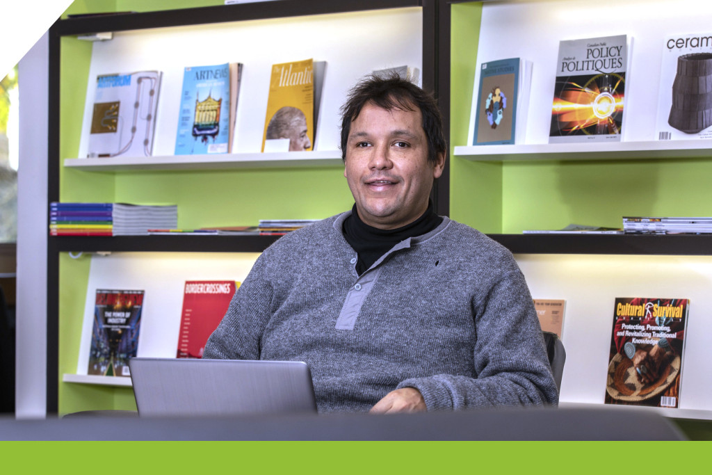 A Indigenous man sitting in front of a bookshelf with a laptop.