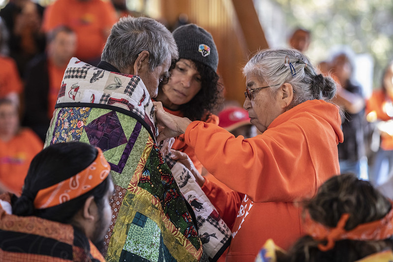 A female elder in an orange shirt wraps a blanket around a male elder. The background is blurred but there is obviously a crowd at an outdoor event.