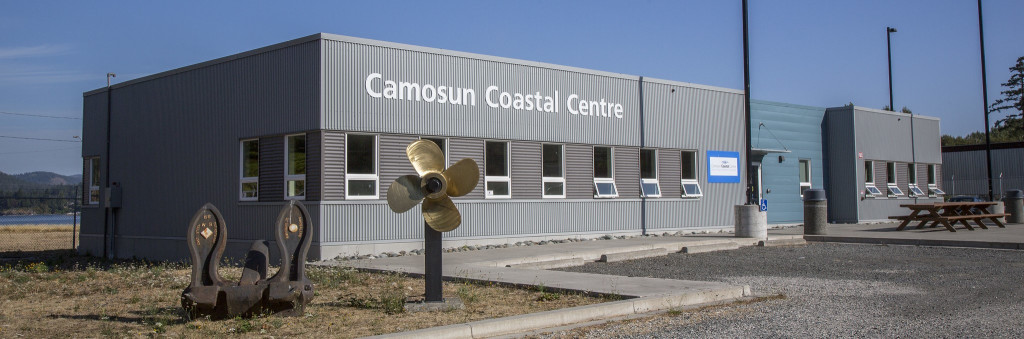An industrial looking building with a the ocean and a shipyard in the background. A large sign over the entrance says sign that says "Camosun Coastal Centre".