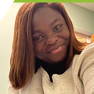 Ayomide smiles at the camera, sitting indoors near a desk with light green walls.