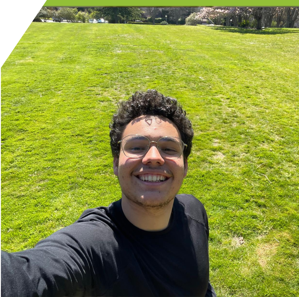 Juan, wearing glasses and a black shirt, smiles as he takes a selfie in a  green grassy field