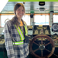 A woman stands at the helm of a large ship or ferry. She has the wheel and is piloting the boat.