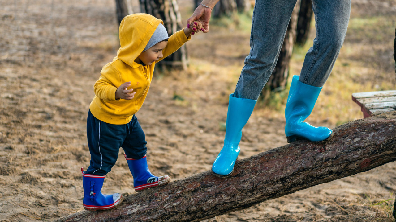 An adult gently leads a toddler across a log on a playground.