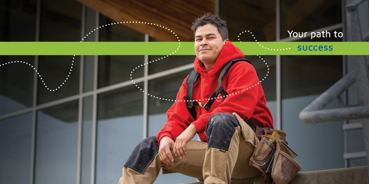 A young indigenous man wearing a red jacket and work person's overalls  and a tool belt sits casually on a ledge, gazing thoughtfully into the distance.