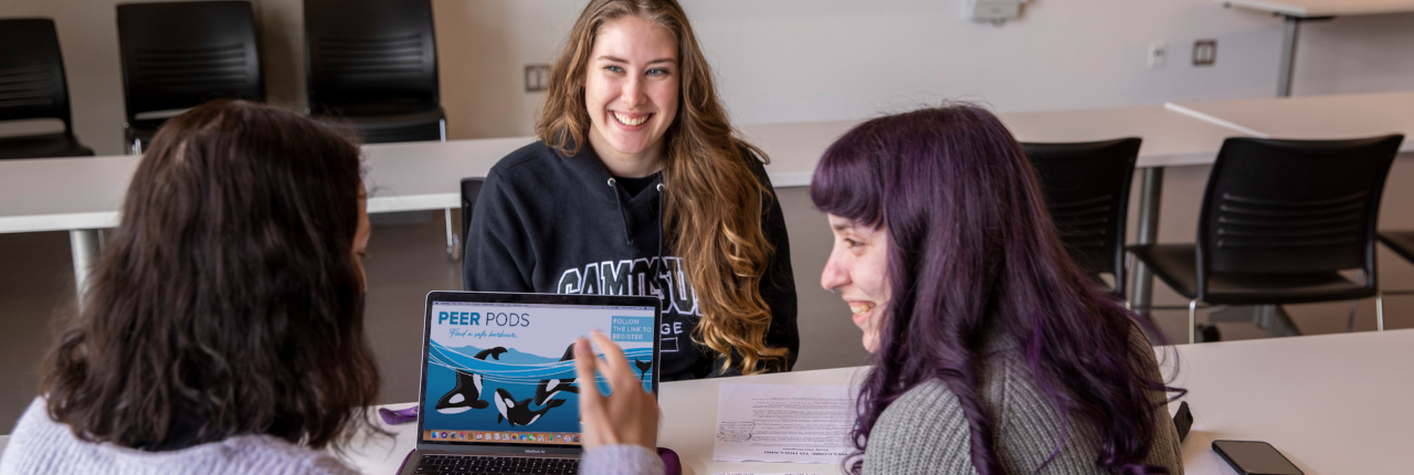 Three students are talking seated in a table where a laptop displays the peer pods logo.