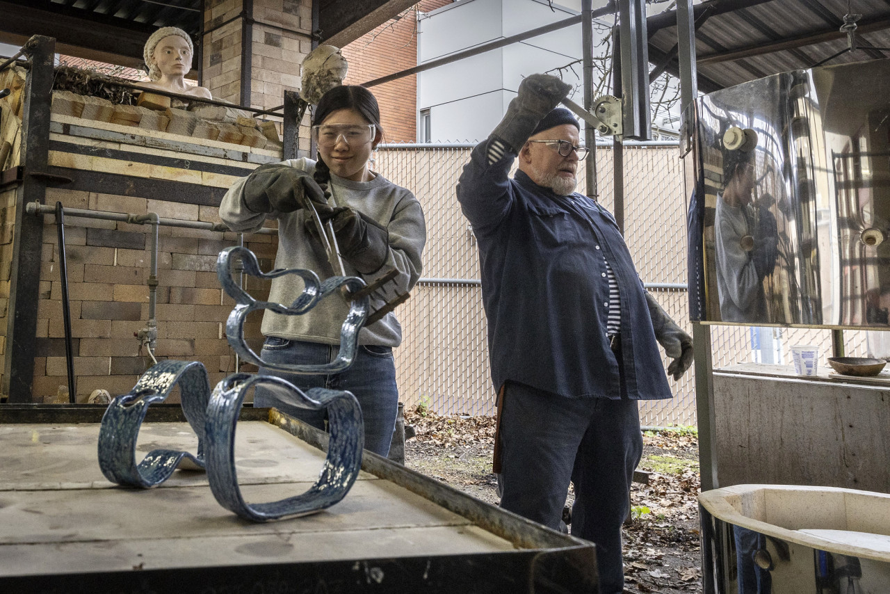 A student uses a set of tongs to set down a sculpture of a Mobius strip on cooling table in front of a ceramics kilim.   