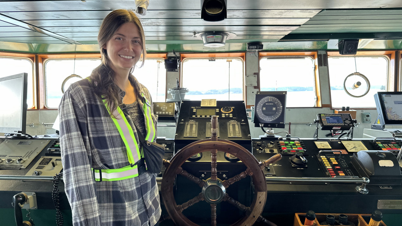 A woman wearing a safety vest stands at the helm of a lager ship or ferry. She is driving the boat.
